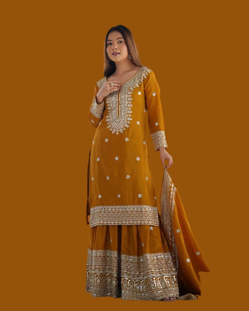 May include: A mustard-coloured salwar kameez outfit with silver embroidery. The long-sleeved tunic has detailed embellishments around the neckline and cuffs, paired with wide-legged trousers and a matching dupatta. The outfit is decorated with small, scattered embellishments.