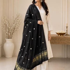 May include: A white long-sleeved tunic and matching trousers paired with a black dupatta. The dupatta features scattered circular silver mirror embellishments and a wide gold metallic border at the hem. The tunic has a V-neckline with embroidery.