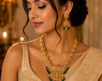 May include: A gold-toned necklace and matching jhumka earrings set featuring intricate metalwork, small green beads, and red accents. The necklace has a central pendant with detailed engravings, while the earrings are bell-shaped with hanging beads.