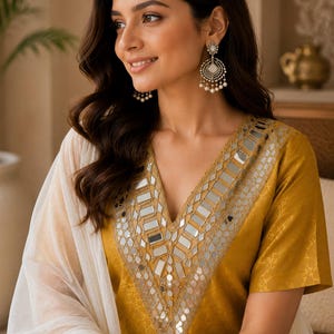 May include: A mustard yellow tunic featuring a V-neckline adorned with intricate silver-toned mirror work embroidery. The garment is paired with a sheer white fabric draped over the shoulder. Silver-toned chandelier earrings with small white beads.