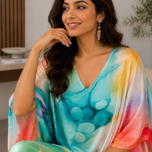 May include: A V-neck kaftan featuring a vibrant watercolor print with blended shades of turquoise, teal, orange, yellow, and pink. The garment has wide, loose sleeves and a smooth, satin-like finish that drapes across the body in a relaxed silhouette.