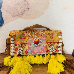 May include: A rectangular, embellished handbag with a brown handle and fringe. The bag features a patchwork design with floral embroidery, cowrie shells, and yellow tassels. The color palette includes shades of yellow, orange, and brown.