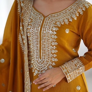 May include: A mustard yellow long-sleeved tunic featuring an intricate silver sequin and thread embroidery pattern around the neckline, cuffs, and along the edge of the matching draped shawl, creating a detailed metallic floral design.