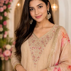 May include: A beige-colored formal dress with intricate embroidery around the neckline and sleeves. The dress features a V-neck design and is paired with a floral-patterned scarf. Gold-colored jewelry, including earrings and bangles, adds a touch of elegance.