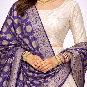 May include: A cream-colored, long-sleeved top and skirt set with gold floral embroidery. A deep purple shawl with gold floral patterns is draped over the shoulders. The model is wearing gold jewelry and colorful bangles.