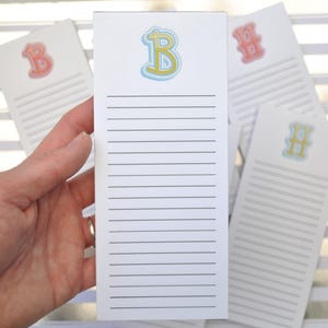 May include: A rectangular white notepad featuring a large letter B in yellow with a blue outline at the top, followed by a series of horizontal black lines for writing. Several other similar notepads with different letters are visible in the background.