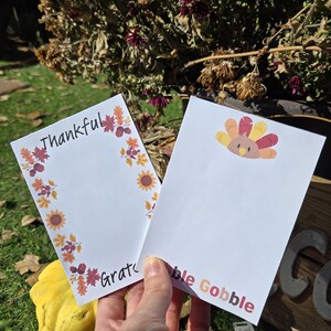 May include: Two white notepads with fall-themed borders. One notepad has a turkey illustration and the text "bble Gobble". The other notepad has a fall leaf border and the text "Thankful" and "Grate".