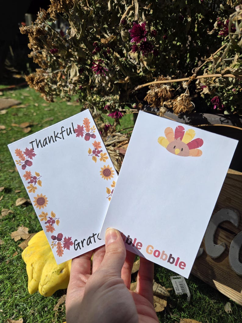 May include: Two white notepads with fall-themed borders. One notepad has a turkey illustration and the text "bble Gobble". The other notepad has a fall leaf border and the text "Thankful" and "Grate".