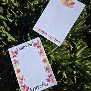 May include: Two blank note cards with fall-themed borders. One card has a turkey illustration and the text "Gobble Gobble". The other card has a border of leaves, sunflowers, and berries and the text "Thankful" and "Grateful".