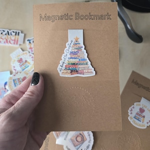 May include: A brown magnetic bookmark with the text "Magnetic Bookmark" printed on it. The bookmark features a colorful stack of books in the shape of a Christmas tree, with the text "all booked for Christmas".