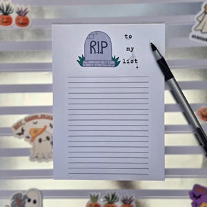 Pode incluir: Um bloco de notas com tema de Halloween com uma ilustra&ccedil;&atilde;o de l&aacute;pide e as palavras "R.I.P." e "to my list". Uma caneta preta e prateada est&aacute; sobre o papel pautado. Adesivos decorativos de fantasmas e ab&oacute;boras est&atilde;o espalhados.