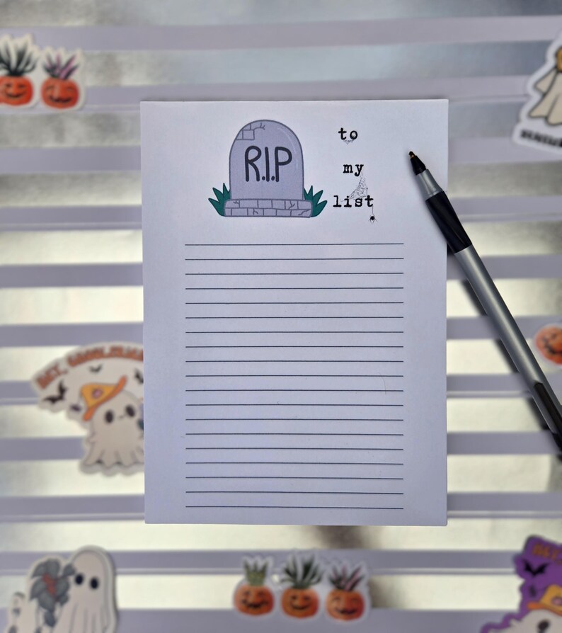Pode incluir: Um bloco de notas com tema de Halloween com uma ilustra&ccedil;&atilde;o de l&aacute;pide e as palavras "R.I.P." e "to my list". Uma caneta preta e prateada est&aacute; sobre o papel pautado. Adesivos decorativos de fantasmas e ab&oacute;boras est&atilde;o espalhados.