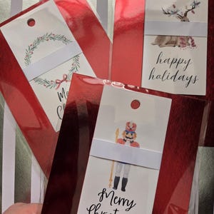 May include: Gift tags in red cellophane packaging. One tag features a wreath design with the text "Merry Christmas." Another says "Happy Holidays" with a deer illustration. A third tag shows a nutcracker with the text "Merry Christmas."