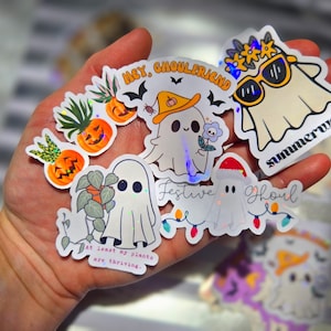 May include: A collection of holographic stickers featuring ghost characters with various themes. Designs include ghosts with sunglasses, hats, and holding plants. Some stickers have text like "Summerween" and "Festive Ghoul".