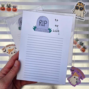 Pode incluir: Um bloco de notas com tema de Halloween com um desenho de l&aacute;pide e as palavras "to my list". O bloco de notas tem p&aacute;ginas pautadas. A imagem inclui adesivos de fantasmas e ab&oacute;boras. O bloco de notas &eacute; segurado por uma m&atilde;o.