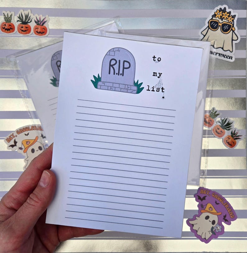 Pode incluir: Um bloco de notas com tema de Halloween com um desenho de l&aacute;pide e as palavras "to my list". O bloco de notas tem p&aacute;ginas pautadas. A imagem inclui adesivos de fantasmas e ab&oacute;boras. O bloco de notas &eacute; segurado por uma m&atilde;o.