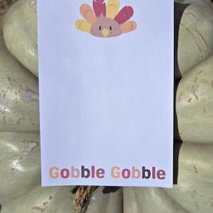 May include: A white card with a colorful turkey illustration and the text "Gobble Gobble" printed in colorful letters. The card is sitting on top of a green pumpkin.
