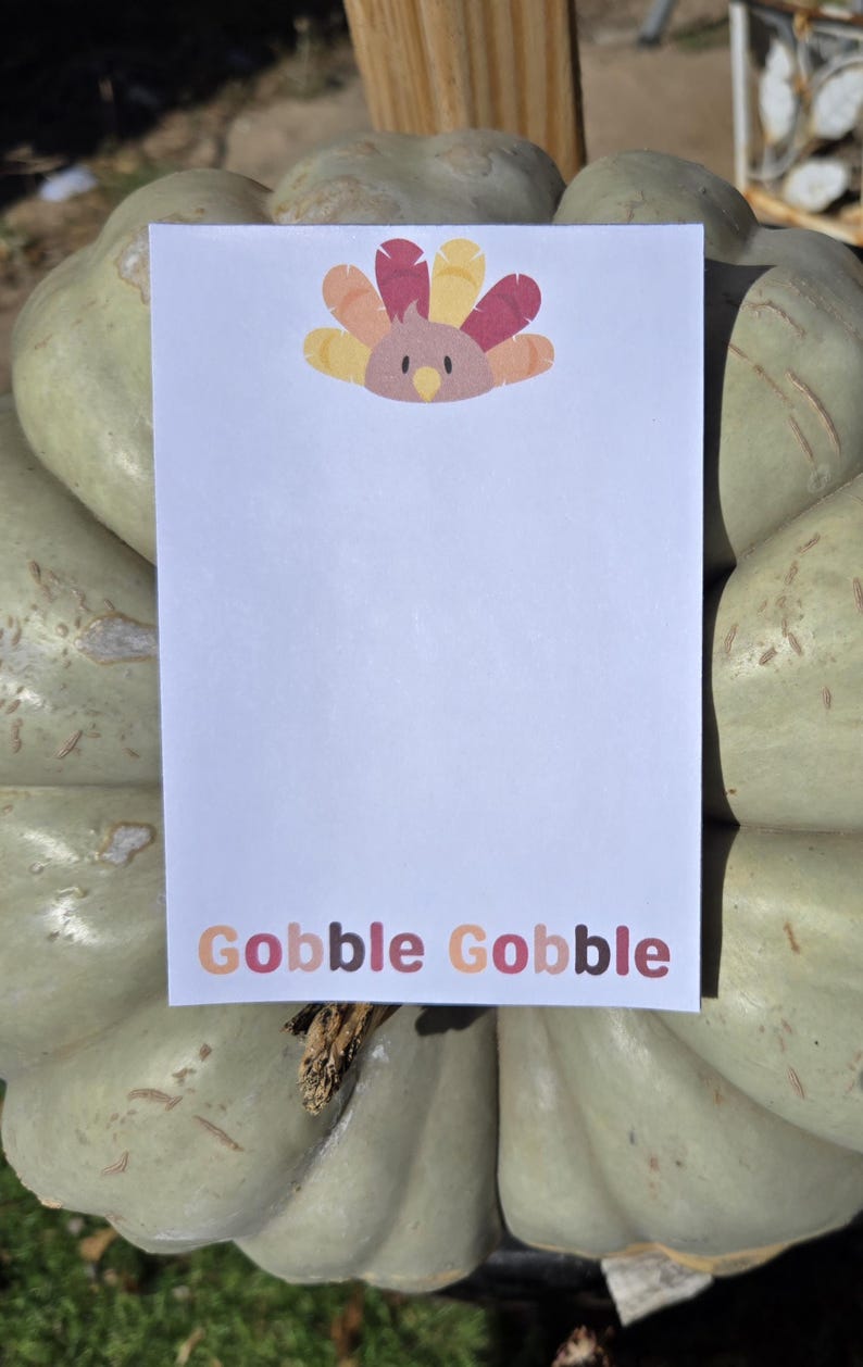 May include: A white card with a colorful turkey illustration and the text "Gobble Gobble" printed in colorful letters. The card is sitting on top of a green pumpkin.