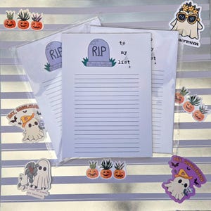 Pode incluir: Blocos de notas pautados com um desenho de l&aacute;pide que diz "R.I.P. to my list". Os blocos de notas s&atilde;o rodeados por autocolantes com tema de Halloween, incluindo fantasmas e h&iacute;bridos de ab&oacute;bora e anan&aacute;s, num fundo listrado prateado e branco.