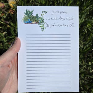 May include: A white lined notepad with a quote about growth and a watercolor illustration of potted plants on a shelf. The quote reads "You are growing, even on the days it feels like you're standing still."