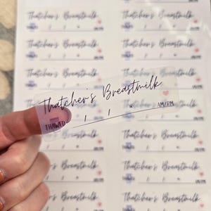 May include: Clear, rectangular labels with the text "Thatcher's Breastmilk" in a handwritten font. The labels include space for AM/PM and a "THAWED" indicator. The background shows more labels.