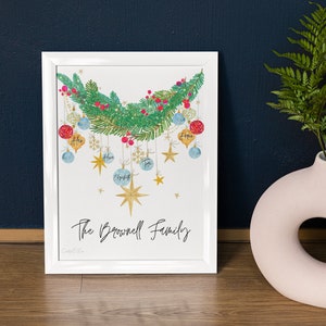 May include: A white framed print with a watercolor illustration of a Christmas garland with ornaments and stars. The ornaments are decorated with the names Alex, Ava, Elizabeth, Tom, and Anna. The text "The Brownell Family" is written below the garland.