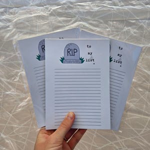 Pode incluir: Uma pilha de blocos de notas pautados com um gr&aacute;fico de l&aacute;pide que diz "R.I.P. to my list". A l&aacute;pide &eacute; cinza com uma base verde. Os blocos de notas s&atilde;o segurados em uma m&atilde;o.