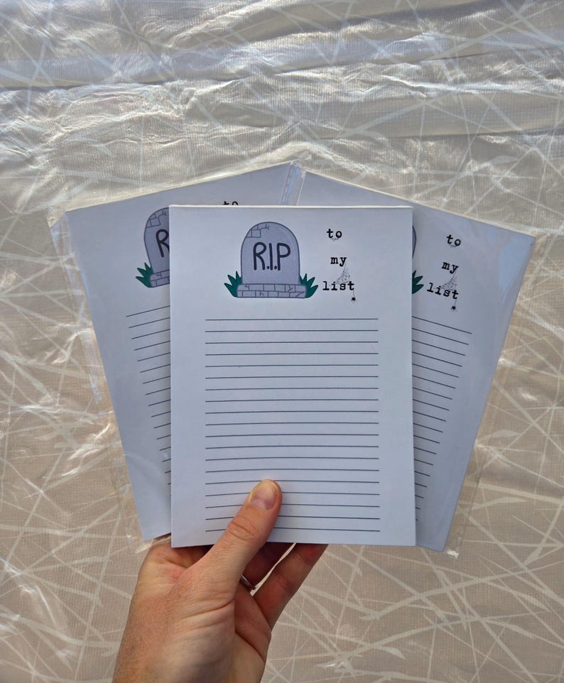 Pode incluir: Uma pilha de blocos de notas pautados com um gr&aacute;fico de l&aacute;pide que diz "R.I.P. to my list". A l&aacute;pide &eacute; cinza com uma base verde. Os blocos de notas s&atilde;o segurados em uma m&atilde;o.