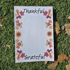 May include: A white card with a fall-themed border featuring sunflowers, leaves, and berries. The card says "Thankful" at the top and "Grateful" at the bottom.