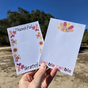 May include: Two white notepads with fall-themed borders. One notepad has the words "Thankful" and "Grateful" written on it. The other notepad has a cartoon turkey and the words "Gobble Gobble" written on it.