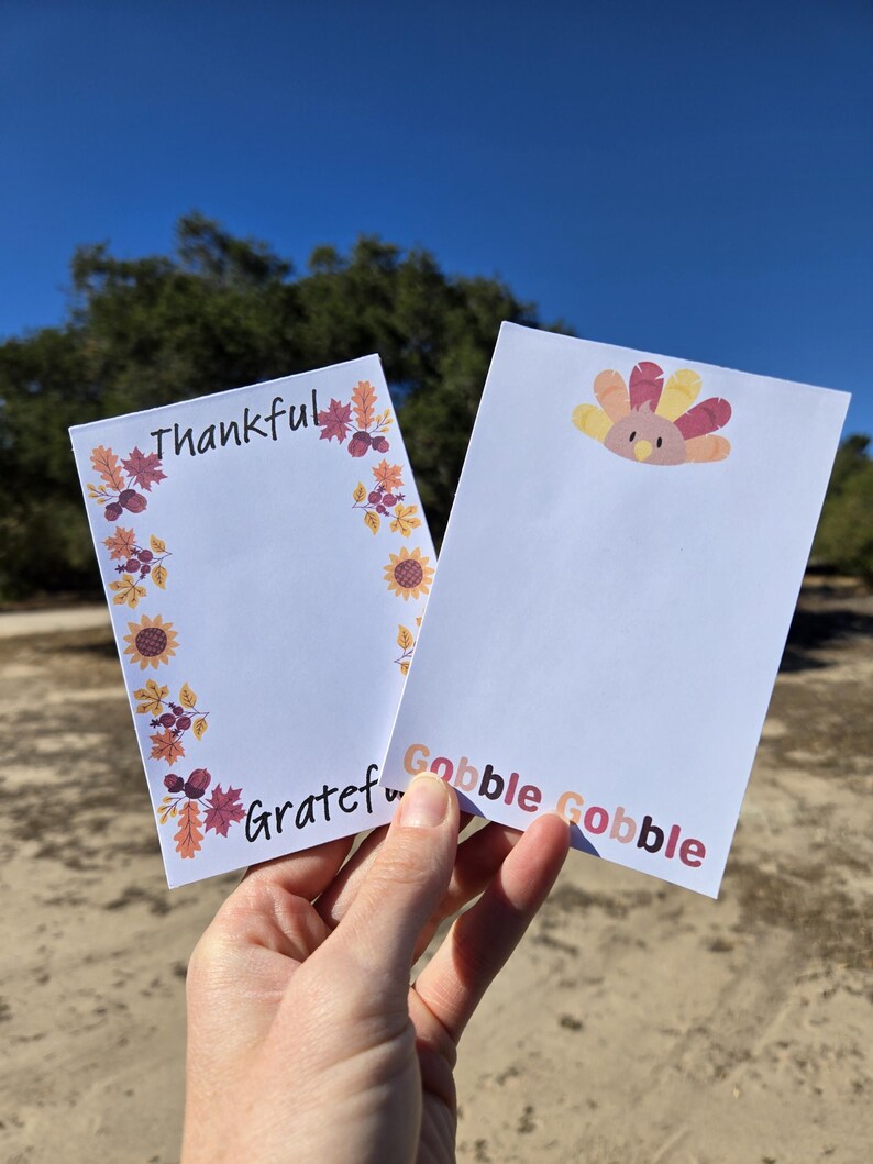 May include: Two white notepads with fall-themed borders. One notepad has the words "Thankful" and "Grateful" written on it. The other notepad has a cartoon turkey and the words "Gobble Gobble" written on it.