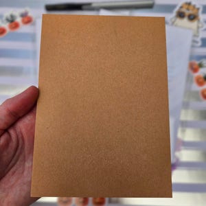 Pode incluir: Um cart&atilde;o retangular castanho com uma superf&iacute;cie texturizada, segurado por uma m&atilde;o. Uma caneta e autocolantes s&atilde;o vis&iacute;veis no fundo.