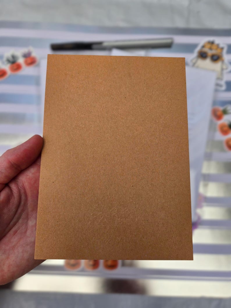 Pode incluir: Um cart&atilde;o retangular castanho com uma superf&iacute;cie texturizada, segurado por uma m&atilde;o. Uma caneta e autocolantes s&atilde;o vis&iacute;veis no fundo.