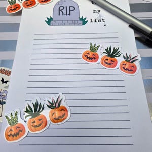 Pode incluir: Um bloco de notas pautado com as palavras "my list" e um gr&aacute;fico de l&aacute;pide que diz "R.I.P." &eacute; decorado com adesivos de ab&oacute;bora laranja com folhas verdes. Uma caneta prateada repousa no bloco de notas.