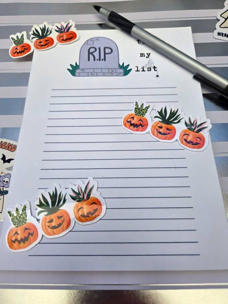 Pode incluir: Um bloco de notas pautado com as palavras "my list" e um gr&aacute;fico de l&aacute;pide que diz "R.I.P." &eacute; decorado com adesivos de ab&oacute;bora laranja com folhas verdes. Uma caneta prateada repousa no bloco de notas.