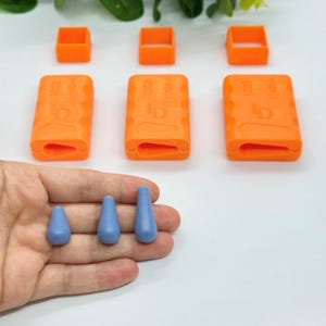 May include: Three orange silicone moulds for making jewellery. The moulds are shaped like a rectangle with a small opening in the centre. There are also three small, light blue silicone teardrop-shaped beads.