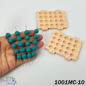 May include: A hand holds strands of turquoise beads on silver pins, next to two beige bead organisers. The beads are round and evenly spaced. The organisers have multiple circular indentations. The text "1001MC-10" is visible.