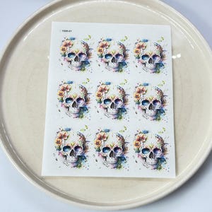 Peut inclure: Une feuille de neuf autocollants de crânes colorés avec des motifs floraux. Les crânes sont blancs avec des yeux noirs et un nez noir. Les fleurs sont dans des tons de rose, violet, bleu, jaune et vert.