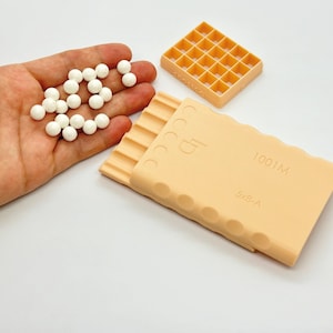 May include: A set of two peach-colored plastic molds for making candy or chocolate. The larger mold has a grid pattern with 20 squares. The smaller mold has a series of ridges and the text "1001M 5x8-A". A hand is holding 20 white round candy pieces.