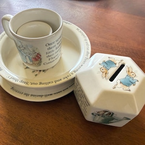 May include: A ceramic Peter Rabbit themed set including a white mug, a shallow bowl, and a hexagonal coin bank. Each piece features illustrations of rabbits in blue jackets and printed text from the story. The bowl and mug have a cream base.