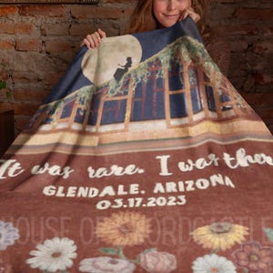 Op de afbeelding: Een bruine fleece deken met een bedrukt beeld van een huis met een silhouet van een persoon op het dak. De tekst "It was rare. I was there. GLENDALE, ARIZONA 03.17.2023" is op de deken gedrukt.