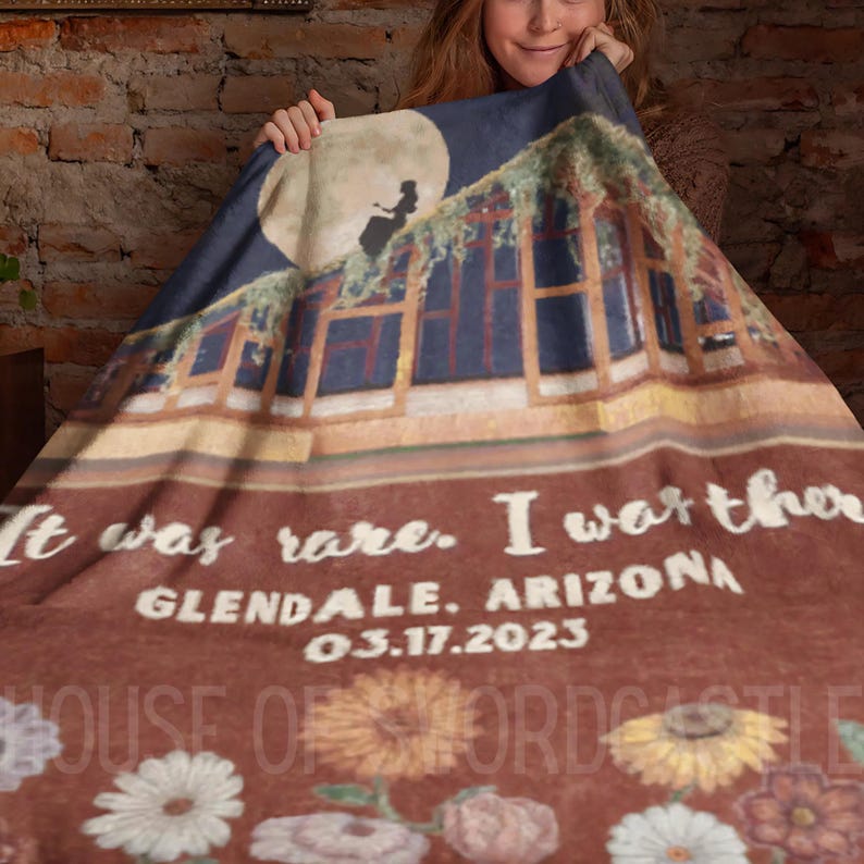 Op de afbeelding: Een bruine fleece deken met een bedrukt beeld van een huis met een silhouet van een persoon op het dak. De tekst "It was rare. I was there. GLENDALE, ARIZONA 03.17.2023" is op de deken gedrukt.