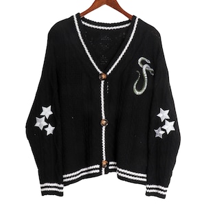 May include: Black knitted cardigan with white trim, featuring a snake and star embroidery on the sleeves. The cardigan has a v-neck and button closure.