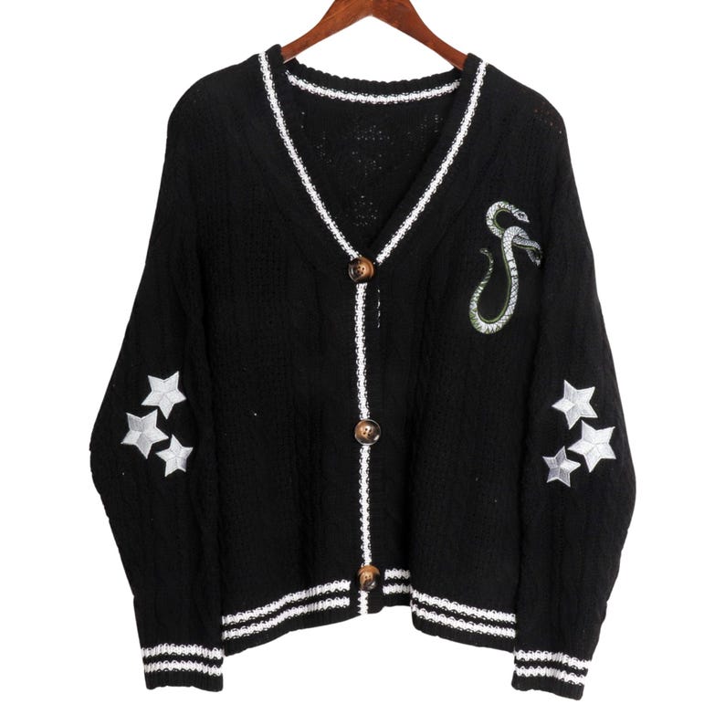May include: Black knitted cardigan with white trim, featuring a snake and star embroidery on the sleeves. The cardigan has a v-neck and button closure.