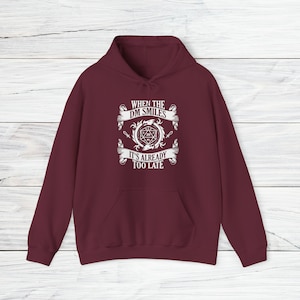 Op de afbeelding: Bordeauxkleurige hoodie met een witte afbeelding met de tekst "When the DM smiles it's already too late". De afbeelding bevat ook een 20-zijdige dobbelsteen en andere fantasy-afbeeldingen.