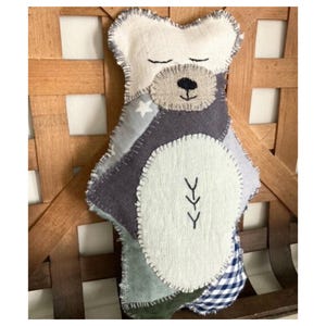 May include: A patchwork stuffed bear toy with a white face, gray body, and blue and green accents. The bear has a stitched-on nose and eyes and a simple embroidered design on its belly.
