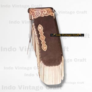 Può includere: Un paio di chaps in pelle marrone con decorazioni floreali e frange. I chaps hanno frange marroni chiare lungo i lati e il fondo. La parte superiore dei chaps ha un motivo floreale decorativo.
