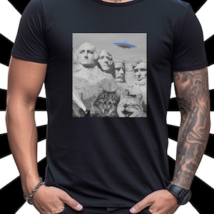May include: Black t-shirt featuring a graphic of Mount Rushmore with cats and a UFO. The image is in grayscale, with the exception of the blue and orange UFO. The shirt has a crew neck and short sleeves.