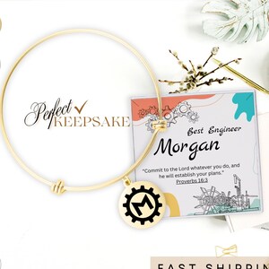 May include: Gold expandable bracelet with a charm featuring a gear with the letter M inside. The charm is attached to a small card with the text "Best Engineer Morgan" and a Bible verse from Proverbs 16:3.