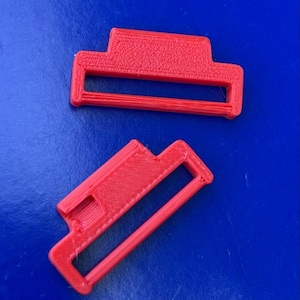 Puede incluir: Dos ajustadores de correa de plástico rojo brillante con un marco rectangular y una barra horizontal central. Estos componentes presentan un acabado superficial texturizado consistente con artículos impresos en 3D, con capas visibles.